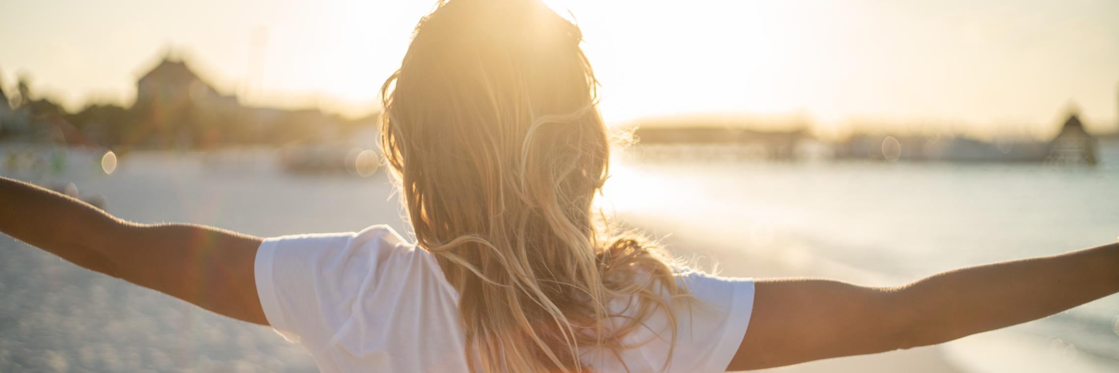 Junge Frau steht am Strand mit ausgestreckten Armen und genießt den Sonnenaufgang.